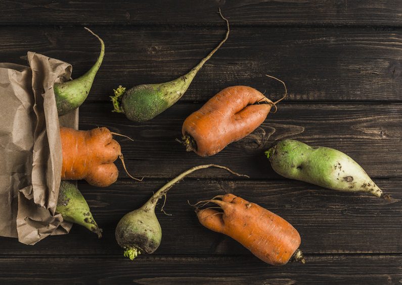 Ugly vegetable green radish and carrot with sprouts on a dark wooden natural background out of a paper bag