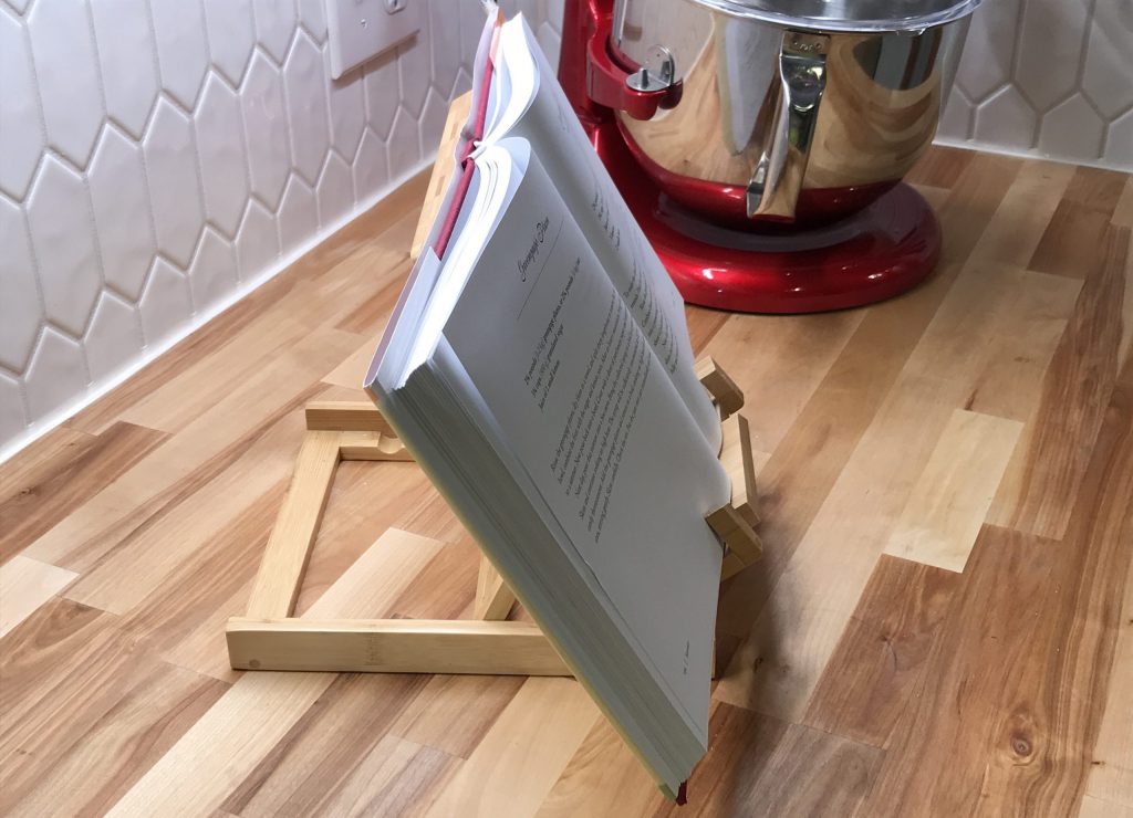 An open cookbook rests in a bamboo holder on a butcher block countertop. A red KitchenAid stand mixer is in the background.