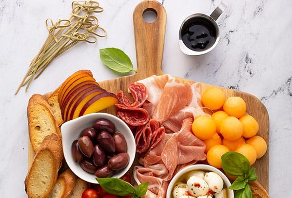 Overhead shot of a portion of a wooden serving board with handle topped with bowls of olives and cheese, plus melon balls, nectarine slices, folded salami and proscuitto slices, toasted baguette slices, and basil leaves for garnish. Bamboo skewers are off to the left side of the board, which rests on a marble surface. A small pitcher of sauce is on the right.