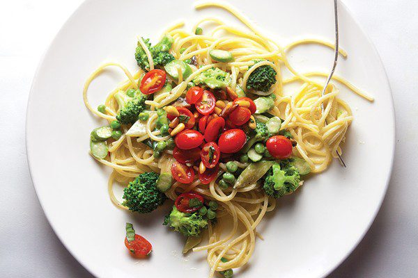 A jumble of vegetables including broccoli, peas, and halved grape tomatoes rests atop of lightly sauced spaghetti noodles on a plain round white plate.