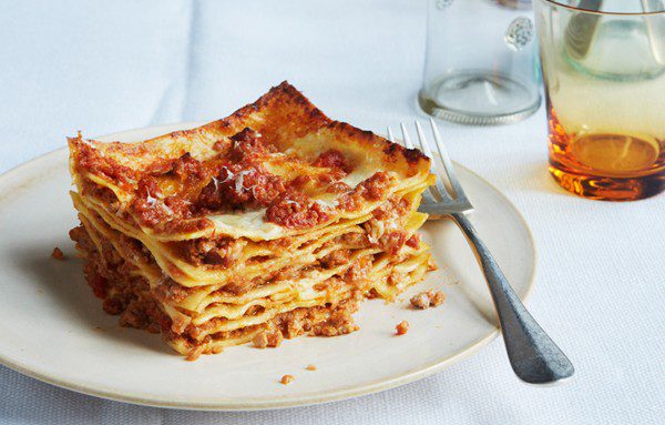 a square of traditional lasagna bolognese on an off-white plate. A fork rests to the right of the lasagna.