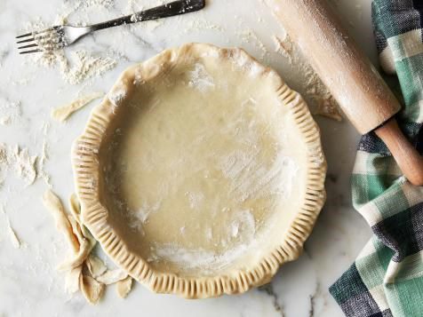 An unbaked pie crust sits in a pie plate with about three-quarters of the edge crimped with a fork. A fork, a rolling pin, and scraps of pie dough surround the crust