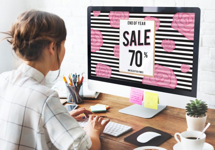 A woman with her hair in a bun sits at a desk in front of a computer screen that has a "sale 70% off" image on it