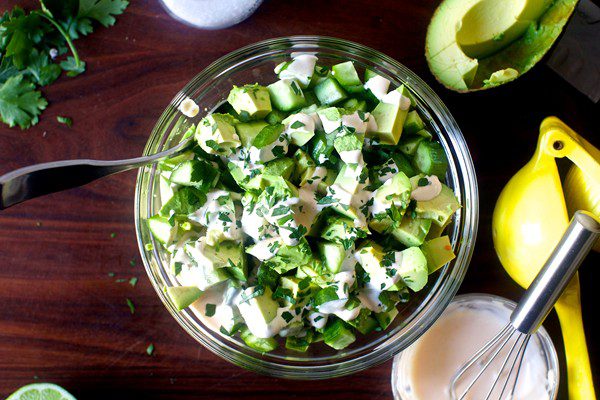 A glass bowl containing a salad with chunks of avocado and cucumber, sprinkled with chopped herbs.