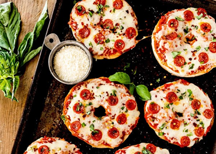 four pepperoni pizza bagels are shown on a dark baking sheet. Basil leaves sit to the left of the image, and a small metal measuring cup placed next to the bagels holds grated cheese.