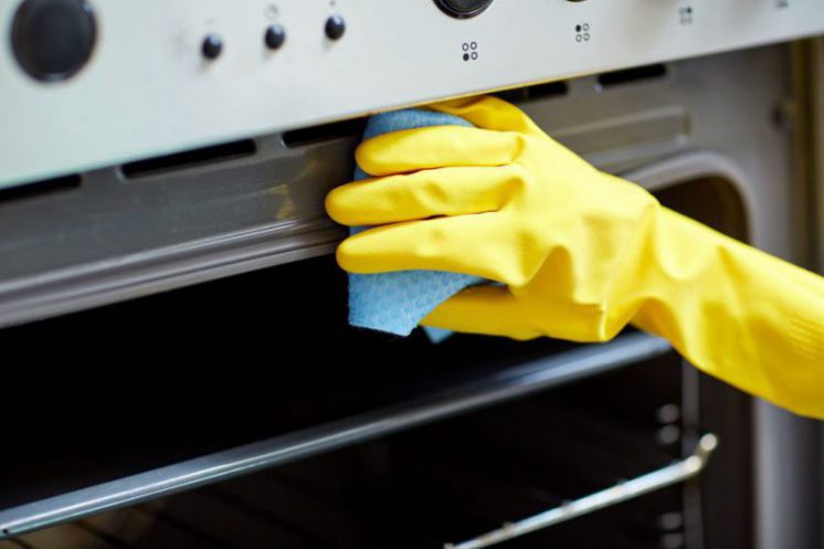 A hand in a yellow cleaning glove wipes across the face of an oven