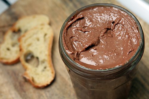 Closeup of a jar of chocolate hazelnut spread with two slices of artisan bread out of focus to the left.
